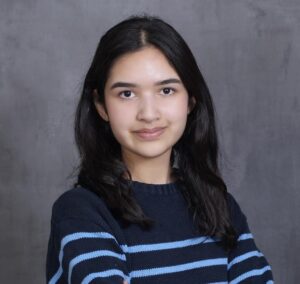 A young woman with dark brown hair wearing a striped shirt sits against a gray background.