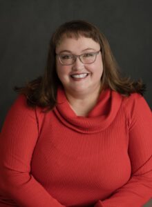 A large woman with brown hair and wearing a red turtle neck smiles at the camera