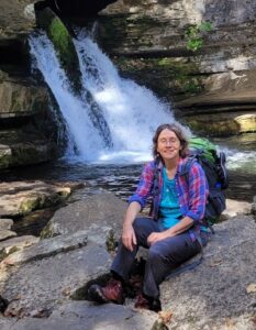 A woman wearing a purple jacket is sitting on a rock in front of a waterfall.