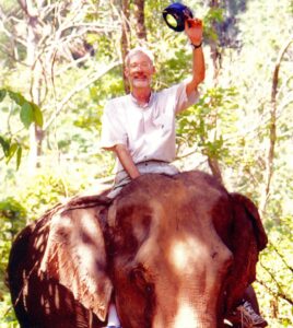 A lean older man waves from the top of an elephant.